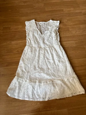Vit broderad sommarklänning - Söt vit klänning i bomull med broderade spetsdetaljer och volang nertill. Klänningen är ärmlös med v-ringning och har ett luftigt, romantiskt uttryck. Perfekt för varma dagar och somriga tillfällen.