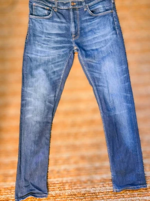 Blå slim fit jeans från Nudie Jeans - Säljer ett par klassiska blå slim fit jeans från Nudie Jeans. Jeansen har fem fickor, snygga slitningar och kontrastsömmar. Tillverkade i mjuk denim med stretch för extra komfort. Perfekta till en avslappnad stil.