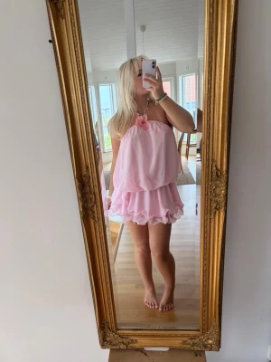Rosa volangklänning med blomdetalj - Supersöt rosa kortklänning med volanger och en stor blomma framtill vid halsen. Klänningen har tunna axelband och en luftig, flowy passform som ger en romantisk vibe. Perfekt för varma dagar och somriga tillfällen.