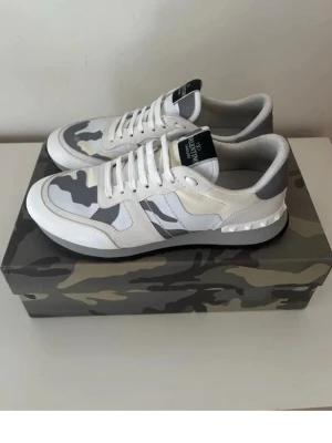 Valentino sneakers med camomönster - Snygga sneakers från Valentino i vitt och grått med camouflagemönster. Skorna har vita snören, svart och vit sula samt detaljer i läder och textil. Märkeslogga på plösen och cool design som sticker ut. Priset är ej hugget i sten. Hör av er för frågor 