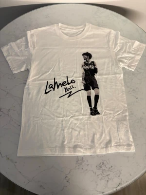 Vit LaMelo Ball t-shirt med tryck - Snygg vit t-shirt med svart tryck av LaMelo Ball och hans signatur. T-shirten har klassisk passform och korta ärmar. Perfekt för dig som gillar basket och streetwear.