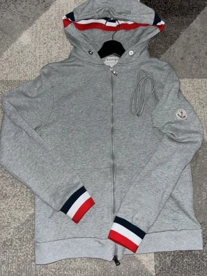 Moncler hoodie  - Snygg grå hoodie från Moncler med dragkedja framtill och huva. Detaljer i rött, vitt och blått på muddar och huva. Moncler-logga på ärmen och praktiska snören i huvan. Tillverkad i mjuk bomullsblandning för skön känsla.