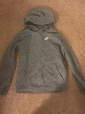 Grå hoodie från Nike med logga - Snygg grå hoodie från Nike med klassisk vit logga på bröstet. Hoodien har känguruficka framtill och är tillverkad i mjukt material som passar perfekt till en avslappnad stil. Långärmad med huva och ribbade muddar.