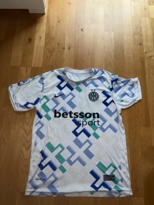 Inter Milan bortatröja Nike Lautaro - Säljer en Inter Milan bortatröja från Nike med LAUTARO och nummer 10 på ryggen. Tröjan är vit med blå och gröna geometriska mönster, har korta ärmar och klubbens logga på bröstet. Perfekt för fotbollsfans som vill sticka ut på planen.