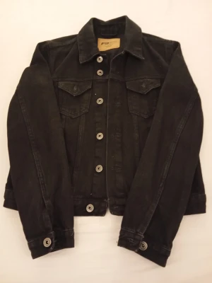 Svart jeansjacka från Fb Sister  - Stilren svart jeansjacka från Fb Sister med klassisk krage, silverfärgade knappar och två bröstfickor med lock. Jackan har raka ärmar och en tidlös design som passar till många olika outfits.
