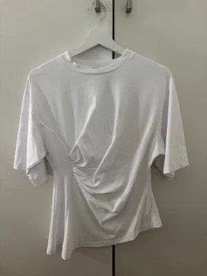 Vit t-shirt med detalj - Säljer en vit t-shirt med korta ärmar och unik detalj på sidan som ger en snygg twist. T-shirten har rund halsringning och är gjord i mjukt bomullstyg. Perfekt för dig som gillar stilrena plagg med en cool detalj. Aldrig använd!