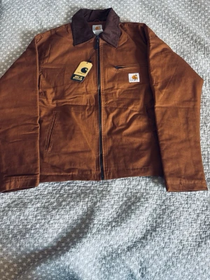 Brun workwear jacka från Carhartt - Snygg brun workwear jacka från Carhartt med dragkedja framtill, mörkbrun krage och klassisk logotyp på bröstet. Jackan har två sidofickor och en bröstficka med dragkedja. Tillverkad i slitstark bomullscanvas, perfekt för en streetwear-look.