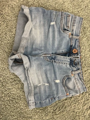 Blå jeansshorts från fb  Sister - Säljer ett par ljusblå jeansshorts från fb Sister med klassisk femficksdesign, uppvikta benslut och slitna detaljer framtill. Shortsen har knapp och dragkedja i gylfen samt läderpatch med logga bak. Perfekta för varma dagar!