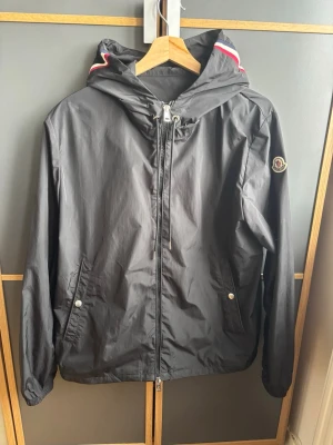 Svart Moncler vindjacka med huva - Svart vindjacka från Moncler med huva och dragkedja framtill. Jackan har två fickor med tryckknappar och Moncler-logga på ärmen. Röd och vit detalj på huvan ger en sportig touch. Perfekt för blåsiga dagar.