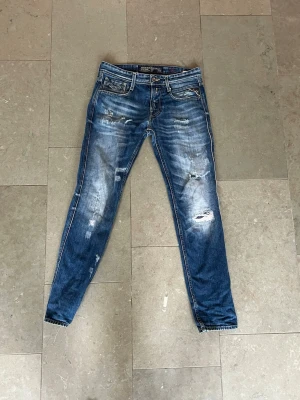 Blå slitna skinny jeans med detaljer - Säljer ett par blå skinny jeans med slitningar och distressed detaljer framtill och baktill. Jeansen har klassisk femficksdesign, låg midja och dragkedjegylf. Perfekta för en avslappnad och trendig look.