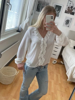 Vit blus med volangdetaljer - Snygg vit blus med djup V-ringning och volang längs halsringningen och framtill. Lätt och luftig modell med långa ärmar och diskreta knappar. 