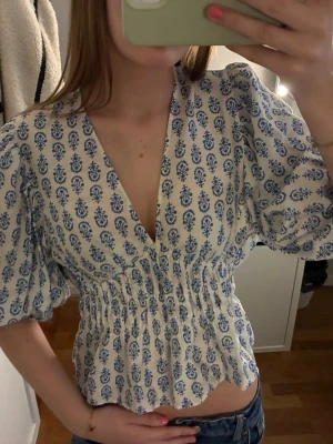 Vit blus med blått mönster - Söt vit blus med blått blommigt mönster och djup v-ringning. Blusen har korta puffärmar och smockad midja som ger en snygg siluett. Perfekt till jeans eller kjol för en trendig look.