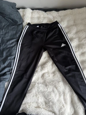 Svarta adidas  byxor med vita rän - Svarta adidas byxor   med vita ränder längs sidorna och elastisk midja. Byxorna har en sportig look och är tillverkade i ett glansigt, mjukt material. Perfekta för en avslappnad stil eller träning.