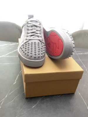 Gråa Louboutin sneakers med nitar - Säljer ett par gråa sneakers från Christian Louboutin med ikoniska röda sulor och coola nitar över hela ovandelen. Skorna har snörning och är tillverkade i mocka för en lyxig känsla. Perfekta för dig som vill sticka ut med din stil.