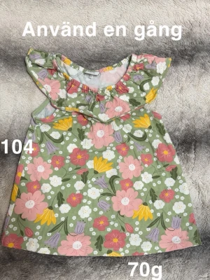 Blommig topp med volangkrage - Söt kort topp med blommigt mönster i rosa, gult, vitt och lila på grön botten. Volangkrage och ärmlös design ger en lekfull känsla. Perfekt för varma dagar och somriga tillfällen.🌸 Använd en gång. Storlek 104