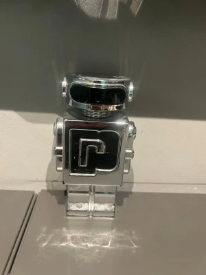 Paco Rabanne Phantom parfym robotflaska - Cool parfymflaska från Paco Rabanne i form av en robot. Flaskan är helt i blankt silverfärgat material med svart detalj på framsidan och ett stort P. Perfekt som statement-piece på parfymhyllan. Kommer utan box och är typ 70 ml kvar.