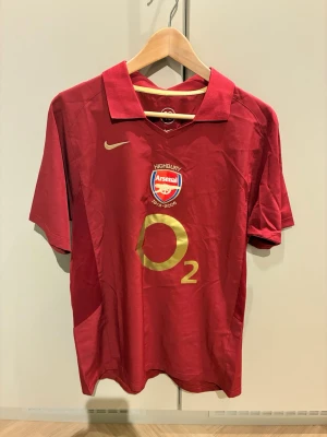 Arsenal Highbury tröja Pires #7 - Röd Arsenal Highbury 1913-2006 fotbollströja från Nike med guldfärgade detaljer, O2-logga och klubbmärke framtill. På ryggen står det 'Pires' och nummer 7 i guld. Klassisk pikékrage och korta ärmar. Perfekt för fotbollsfans!