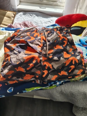 Camo badbyxor i orange och svart - Säljer ett par badbyxor med camomönster i orange, svart och grått. De har elastisk midja med dragsko och en bakficka. Perfekta för stranden eller poolen. Materialet känns lätt och snabbtorkande.