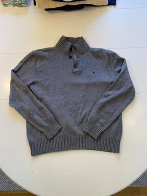Polo by Ralph Lauren grå tröja, storlek M - Grå stickad tröja med hög krage och knappslå. Tröjan har en broderad logotyp på bröstet.  Tröjan har någon typ av svart sträck på baksidan som man knappt tänker på i verkligheten. Har inte testat att tvätta tröjan så kan va så att de försvinner då.   Storlek: M  Längd: 59 cm  Armhålsbredd: 58 cm