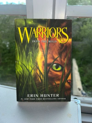 Warriors: Into the Wild av Erin Hunter - En spännande fantasybok på engelska, 'Warriors: Into the Wild' av Erin Hunter. Omslaget har en grön och gul design med en katt som gömmer sig i gräset. Perfekt för dig som gillar äventyr, djur och episka berättelser om klaner och mod.