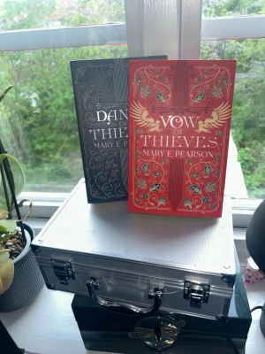 Vow of Thieves & Dance of Thieves - Två snygga inbundna fantasyböcker av Mary E. Pearson: 'Vow of Thieves' i rött med guldiga detaljer och 'Dance of Thieves' i svart med silverdetaljer. Båda har dekorativa omslag med blommönster och är perfekta för dig som gillar spännande äventyr och romantik.