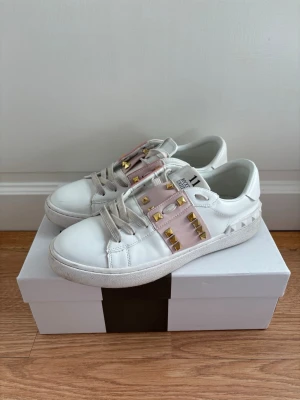 Valentino vita sneakers med nitar - Snygga vita sneakers från Valentino med rosa detaljer och guldiga nitar på sidorna. Skorna har en klassisk låg modell och är tillverkade i läder med vita snören och en chunky sula. Perfekta för dig som vill sticka ut med en lyxig touch.