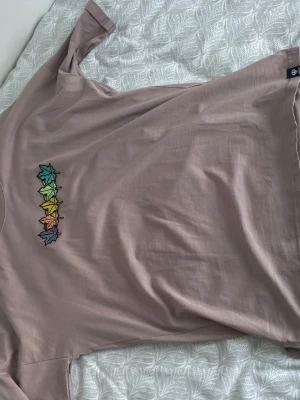 Ljuslila t-shirt med broderade löv - Säljer en ljuslila t-shirt med korta ärmar och broderade löv i regnbågens färger på bröstet. T-shirten är i mjuk bomull och har en avslappnad passform. Perfekt för dig som gillar färgglada detaljer och en chill stil.