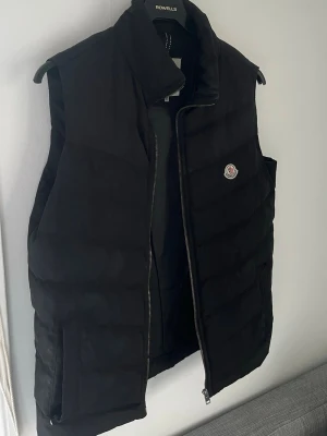 Svart dunväst från Moncler - Snygg svart dunväst från Moncler med klassisk quiltad design och dragkedja framtill. Västen har Monclers ikoniska logotyp på bröstet och hög krage för extra stilpoäng. Perfekt att slänga över en hoodie eller tröja för en trendig look.