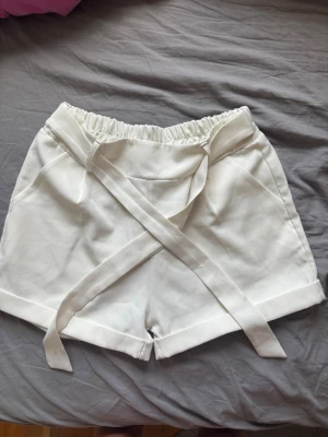 Vita shorts med knytband🫶🏻 - Snygga vita shorts med hög midja och elastisk resår. De har breda ben och ett trendigt knytband framtill som ger en cool touch. Perfekta för varma dagar och lätta att matcha med olika toppar. Vet inte storlek men passa mig som har xs/s. Lite nopprig på rumpan och en liten fläck på bandet🤍