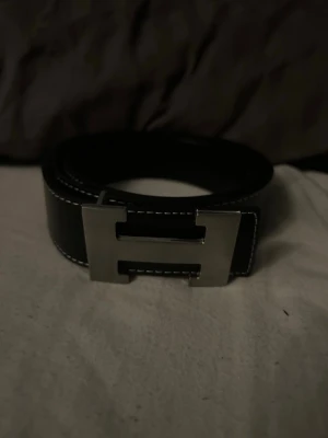 Svart skärp med H-spänne - Stilrent svart bälte med en stor, silverfärgad H-formad metallspänne och vita kontrastsömmar längs kanterna. Perfekt accessoar för att lyfta din outfit med en klassisk och modern touch.