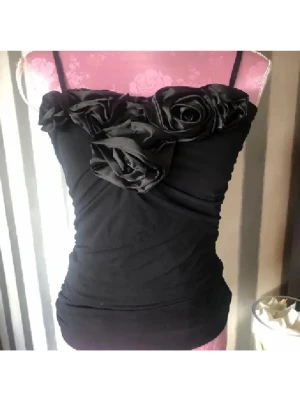 Svart linnetopp med rosdetaljer - Supersnygg svart linnetopp med smala axelband och stora tygrosor längs urringningen. Toppen är figurnära och har en elegant, feminin look. Perfekt för dig som vill sticka ut med coola detaljer och en stilren vibe. Oanvänd 