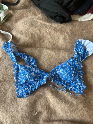 Blå blommig bikiniöverdel med volang - Supersöt blå bikiniöverdel med vitt blommönster och volangdetaljer längs kanterna. Perfekt för stranddagar och sommarhäng. Justerbara axelband för bästa passform.