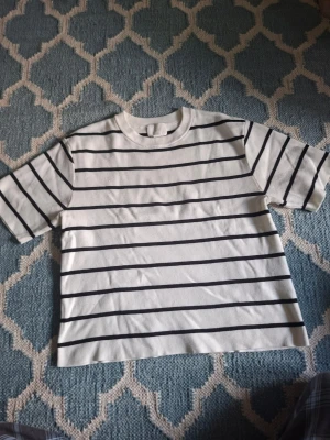 Vit t-shirt med svarta ränder - Säljer en vit t-shirt med svarta horisontella ränder. T-shirten har rund hals och korta ärmar. Perfekt för en avslappnad och stilren look. Materialet känns mjukt och bekvämt, troligtvis bomull.