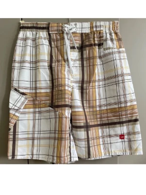 Rutiga cargo shorts med snörning - Säljer ett par rutiga shorts i vitt, brunt och beige med snörning i midjan och praktisk cargoficka på sidan. Shortsen har avslappnad passform och elastisk midja för extra komfort. Perfekta för varma dagar och chill stil.