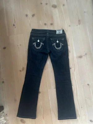 Mörkblå True Religion jeans - Snygga mörkblå jeans från True Religion med utsvängda ben och glitter design. Bakfickorna har märkets ikoniska broderi och detaljer i silver glitter. Jeansen har klassisk femficksdesign och är tillverkade i denim.