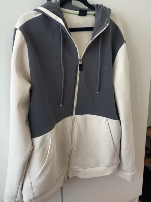 Gråvit hoodie med dragkedja från BOSS - Snygg hoodie från BOSS i grått och vitt med hel dragkedja framtill och två fickor. Hoodien har justerbar huva med snören och en stilren design. Perfekt för en chill och avslappnad look. Materialet känns mjukt och bekvämt mot huden. Kan gå ner i pris vid snabb affär. Skickar samma dag eller dagen efter.