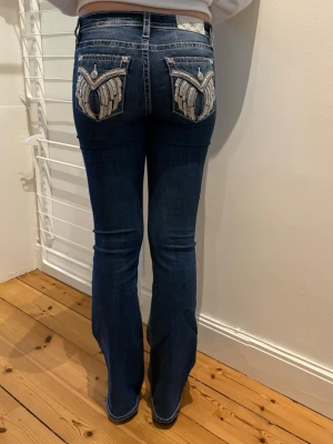 Miss Me jeans. Mid-rise Boot. - Mörkblå bootcut jeans från Miss Me med detaljerade broderier och paljetter på bakfickorna. Jeansen har kontrastsömmar, låg midja och klassisk femficksmodell. Tillverkade i stretchigt denim för skön passform.