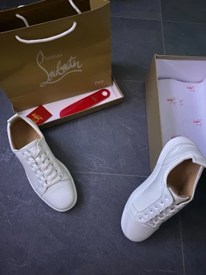 Vita sneakers från Christian Louboutin - Stilrena vita sneakers från Christian Louboutin med klassisk låg silhuett och ikonisk röd sula. Skorna är tillverkade i slätt läder och har vita skosnören samt diskreta detaljer. Perfekta för dig som vill ha en clean och exklusiv look.