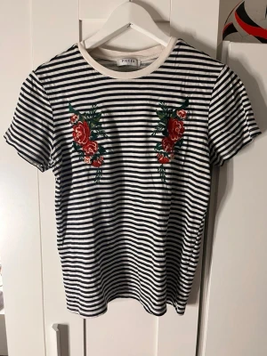 Randig t-shirt med rosbrodyr från Pieces - Svartvit randig t-shirt från Pieces med broderade röda rosor och gröna blad på bröstet. T-shirten har rund halsringning med vit kant och korta ärmar. Den är i storlek L men är passar mer M. 