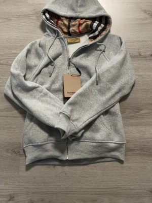 Grå zip-hoodie från Burberry - Snygg grå hoodie med dragkedja från Burberry. Klassisk huva med det ikoniska rutmönstret på insidan, dragsko och ribbade muddar. Perfekt för en chill och stilren look. Materialet är mjukt och skönt, troligtvis bomullsblandning. Passar S också