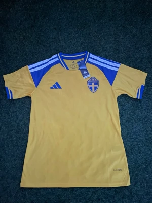 Svensk fotbollströja Adidas gul/blå - Gul och blå fotbollströja från Adidas med Sveriges landslagsemblem på bröstet och klassiska Adidas-ränder på axlarna. Tröjan har rund hals och korta ärmar, perfekt för match eller träning. Materialet är lätt och andas.