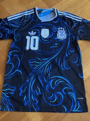 Messi Argentina Adidas fotbollströja blå S - Snygg Argentina fotbollströja från Adidas med blått och svart mönster, nummer 10 och AFA-logga på bröstet. Tröjan har vita detaljer och klassiska Adidas-ränder på axlarna. Tillverkad i lätt och ventilerande material (Climacool). Perfekt för fotboll eller träning.