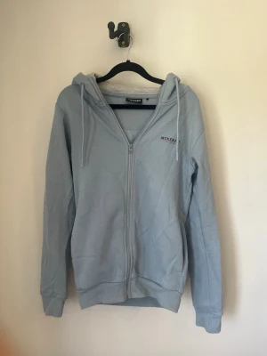 Ljusblå zip-hoodie från McKenzie - Ljusblå hoodie med dragkedja från McKenzie. Klassisk modell med huva och dragsnören, samt ribbade muddar vid ärmslut och nederkant. Liten logga på bröstet. Perfekt för en chill och avslappnad stil. Storlek XS, skick 8/10 