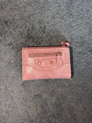 ”City” plånbok - Snygg rosa plånbok i skinn med guldiga detaljer och dragkedja framtill. Plånboken har en unik design med dekorativa nitar och en liten ficka på framsidan. Perfekt för dig som vill ha något stilrent men ändå lekfullt i väskan.