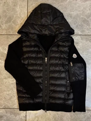 Moncler Cardigan  - Säljer en svart pufferjacka från Moncler med huva och diskret logga på ärmen. Jackan har quiltad design på kroppen och släta svarta ärmar. Perfekt för dig som vill ha en stilren och trendig vinterjacka. NFC funkar