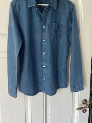 Ljusblå jeansskjorta Tommy Hilfiger - Snygg ljusblå jeansskjorta från Tommy Hilfiger med klassisk krage, bröstficka och vita knappar. Skjortan har lång ärm. Perfekt för en avslappnad och trendig look. Storlek 176 i barn men passar xs-s.
