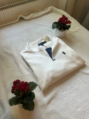 Vit quarter zip från Ralph Lauren - Snygg vit quarter zip-tröja från Ralph Lauren med klassisk broderad logga på bröstet. Tröjan har hög krage och dragkedja framtill. Perfekt för en clean och stilren look. Materialet är mjukt och bekvämt.