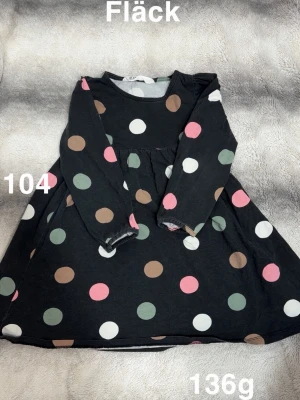 Svart prickig klänning med lång ärm - Svart klänning med färgglada prickar i rosa, brun, grön och vit. Modellen har lång ärm och en lätt utställd kjol. perfekt för dig som gillar lekfulla mönster och bekväma plagg.🌸 Fläck. Storlek 104