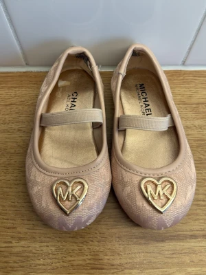 Rosa ballerinaskor från Michael Kors storlek 21  - Snygga ballerinaskor i ljusrosa från Michael Kors med elastiskt band över foten. Skorna har ett hjärtformat guldfärgat MK-emblem framtill och är tillverkade i textil med diskret mönster. Perfekta för dig som gillar stilrena detaljer och trendig design.