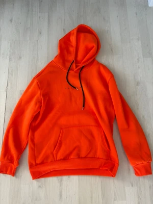 Orange hoodie från Originals - Snygg orange hoodie från Originals med svart dragsko i huvan och tryckt text på bröstet. Hoodien har en stor magficka och ribbade muddar vid ärmslut och nederkant. Perfekt för en avslappnad och cool stil.